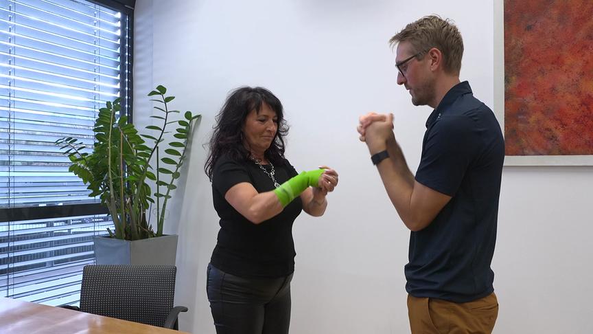 Eine Frau mit einem grünen Verband am Unterarm und ein Sportwissenschafter stehen sich in einem Büro gegenüber und üben eine Bewegungstechnik.