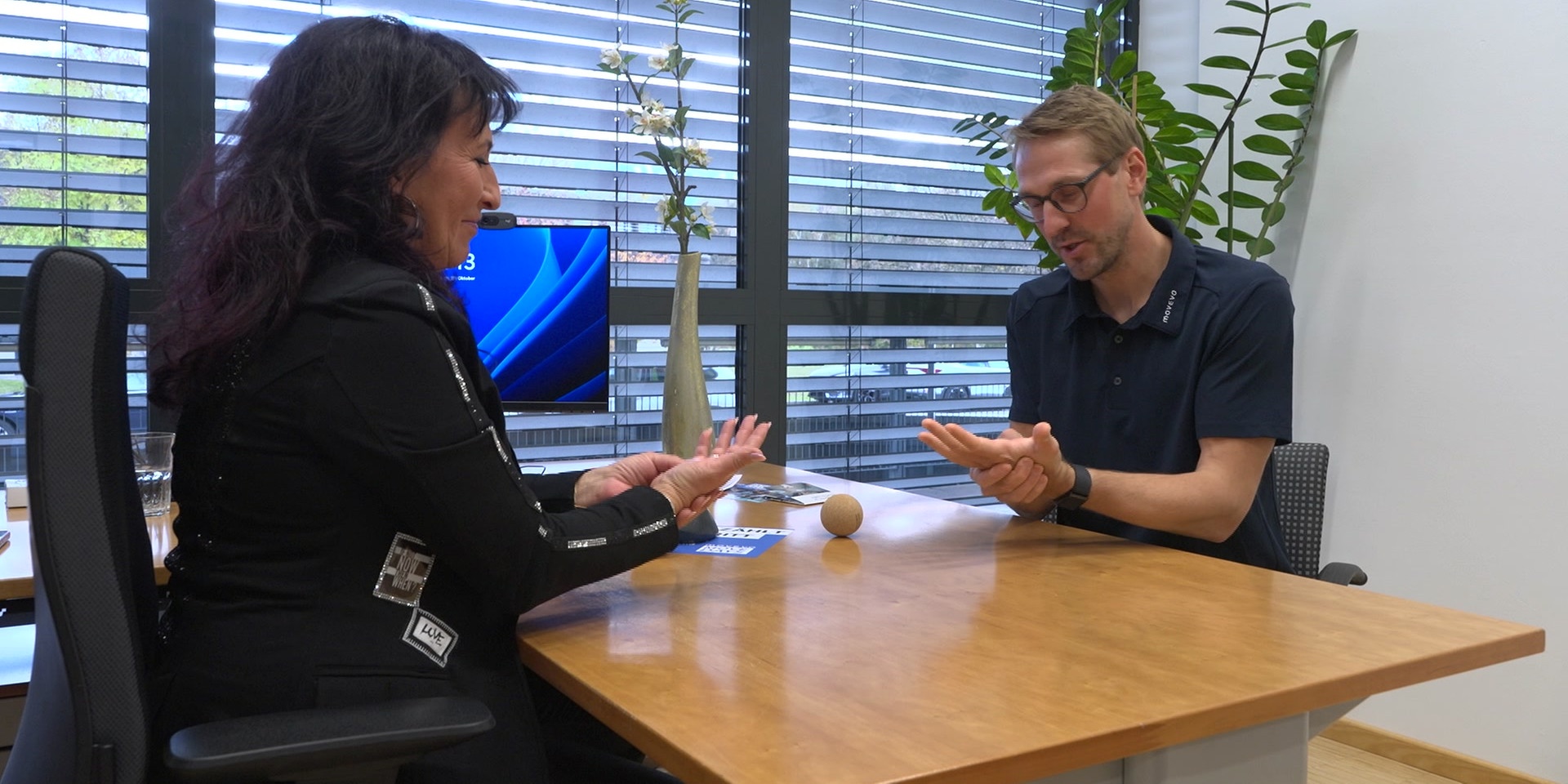 Eine Frau und ein Sportwissenschafter sitzen an einem Schreibtisch und führen eine Unterhaltung mit Gesten, während ein Ball und einige Karten auf dem Tisch liegen.