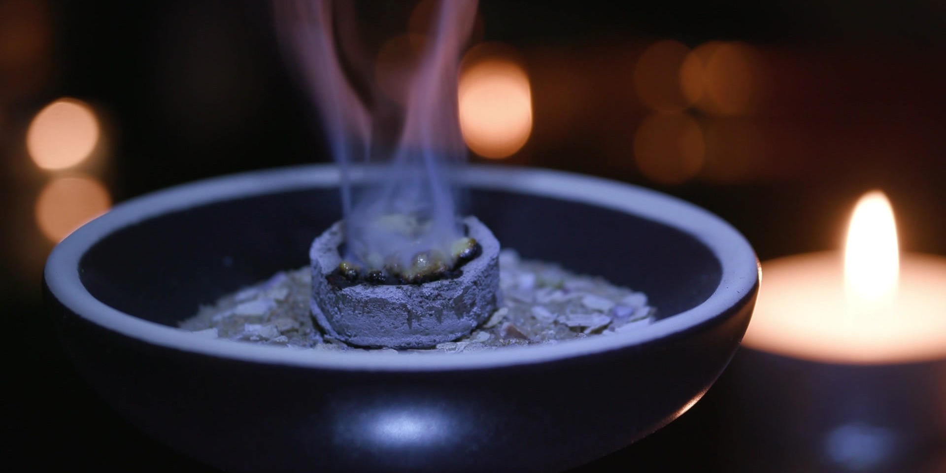 Eine brennende Räucherkohle in einer Schale verströmt duftenden Rauch, umgeben von Kerzenschein.