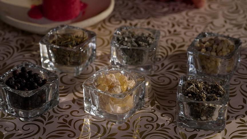 Mehrere kleine Glasgefäße sind mit verschiedenen getrockneten Kräutern und Harzen auf einem verzierten Tisch arrangiert.