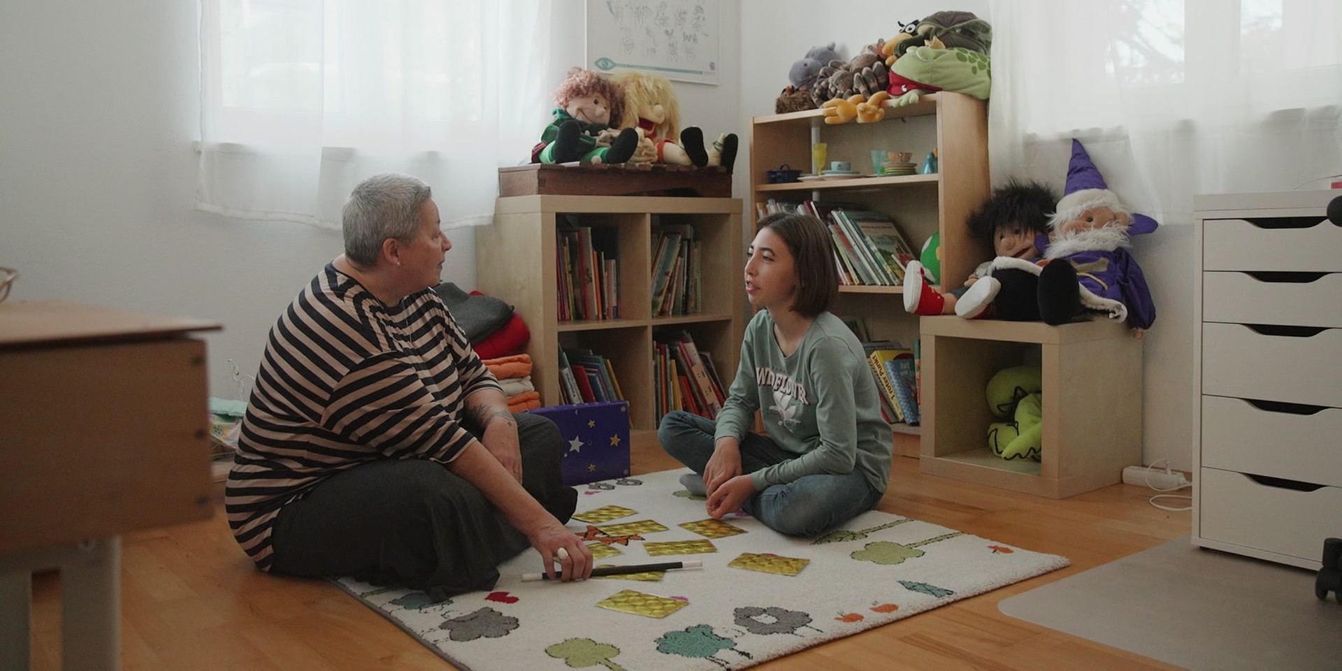 Eine Therapeutin und ein Mädchen sitzen gemeinsam auf einem Teppich in einem gemütlichen Spiel- und Leseraum bei einer therapeutischen Zaubersitzung.