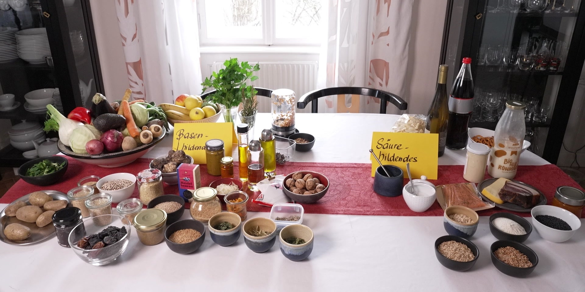 Auf einem Tisch in einem Esszimmer sind zahlreiche Lebensmittel und Zutaten übersichtlich arrangiert und mit gelben Kärtchen als „basenbildend“ und „säurebildend“ gekennzeichnet.
