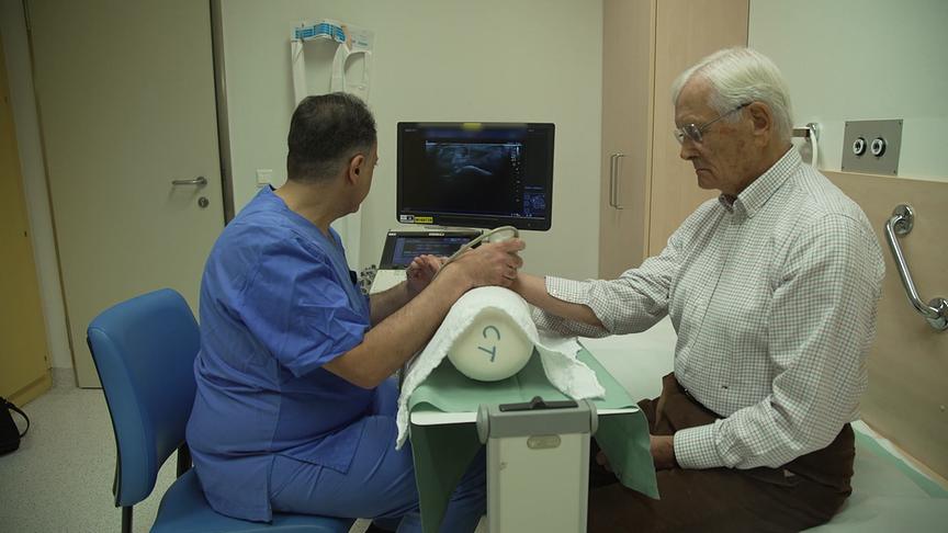 In einem Untersuchungszimmer führt ein Arzt eine Ultraschalluntersuchung am Arm eines sitzenden älteren Mannes durch.