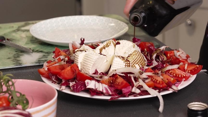 Ein bunter Salat aus Tomaten, Radicchio und Zwiebeln wird mit einem dunklen Öl beträufelt, oben liegt ein großer Rohmilch-Frischkäse.
