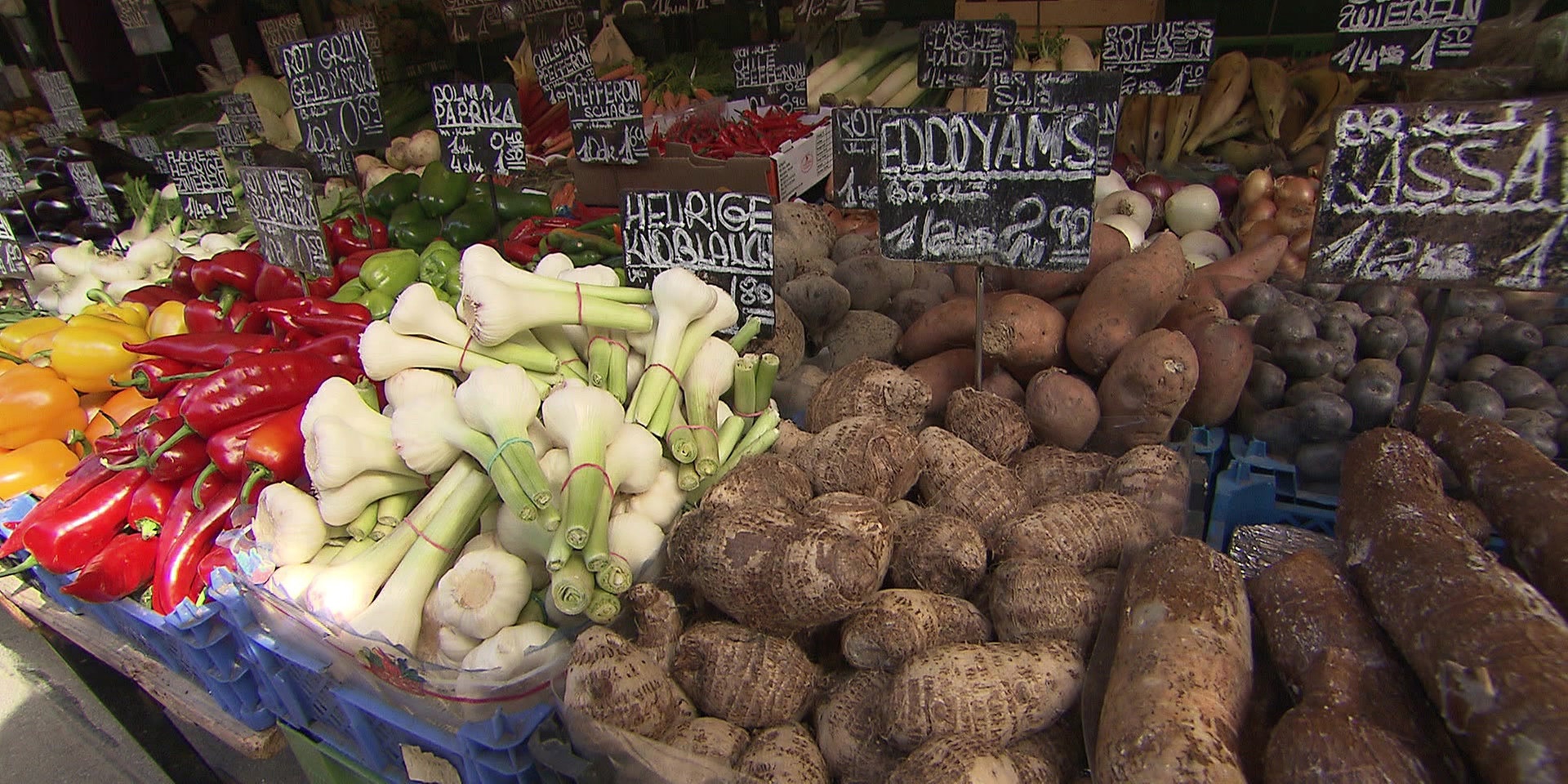 Ein Marktstand zeigt eine große Auswahl an Gemüse wie Paprika, Lauchzwiebeln, Yams und Kartoffeln mit handbeschriebenen Preisschildern.