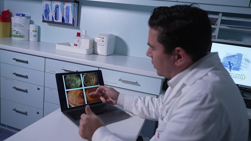 In einem Behandlungsraum zeigt ein Arzt auf einem Laptop endoskopische Aufnahmen einer Darmspiegelung und erklärt diese.