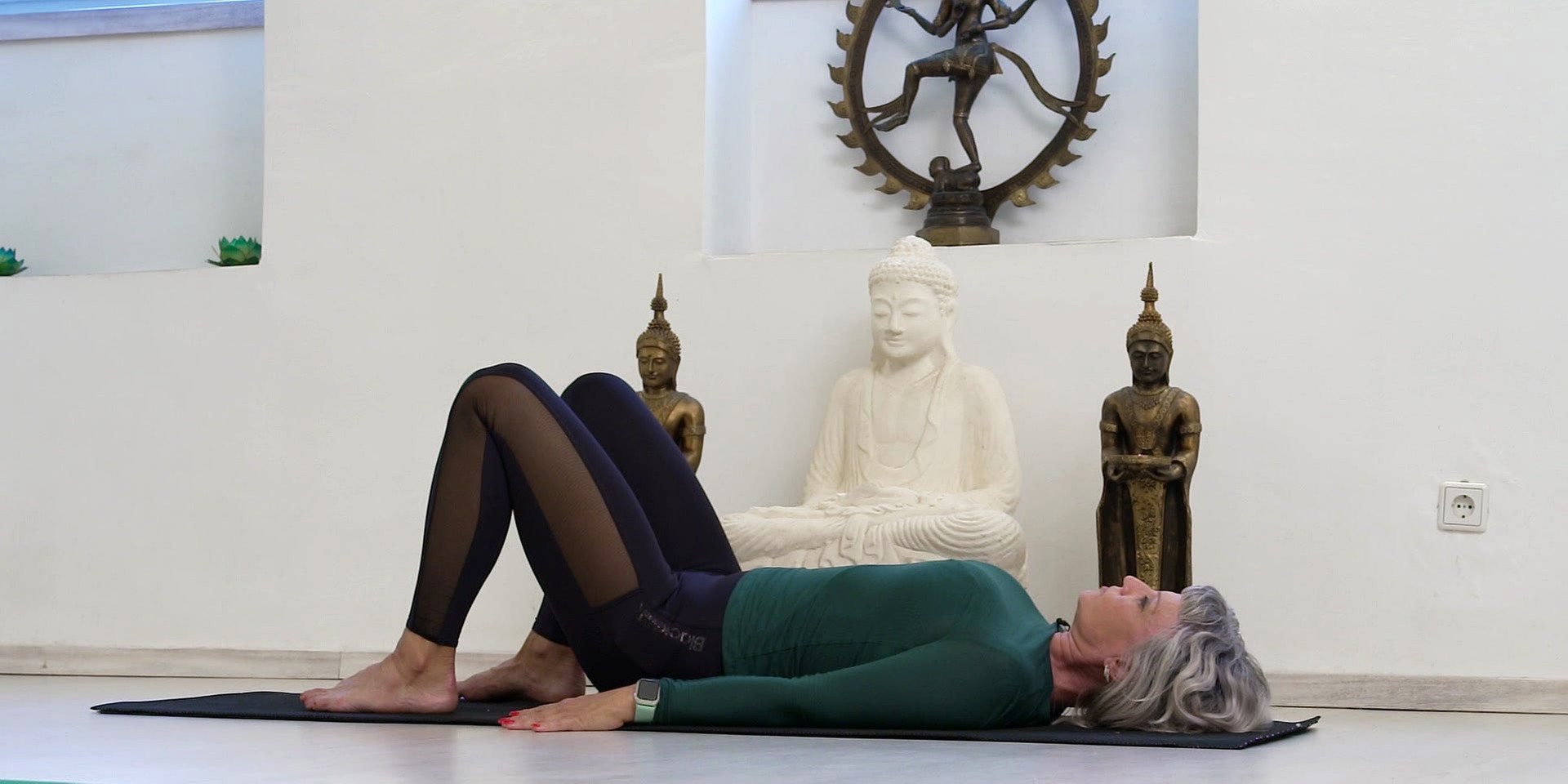 Eine Frau liegt auf einer Yogamatte in Rückenlage mit aufgestellten Beinen in einem ruhigen Raum mit Buddha‑Statuen im Hintergrund.