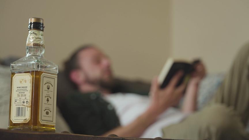 Eine Person liegt entspannt auf einem Sofa und liest ein Buch, im Vordergrund steht eine Whiskeyflasche.