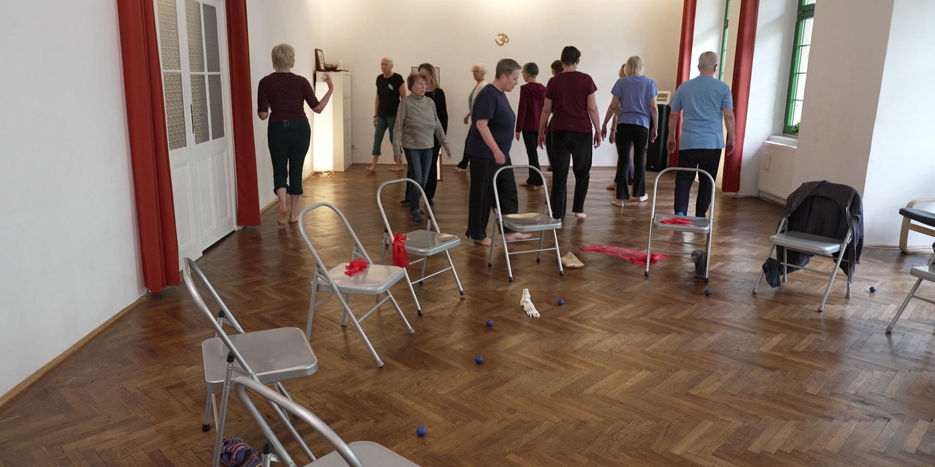 Eine Gruppe von Menschen geht barfuß in einem Kursraum zwischen aufgestellten Stühlen und Trainingsmaterial.