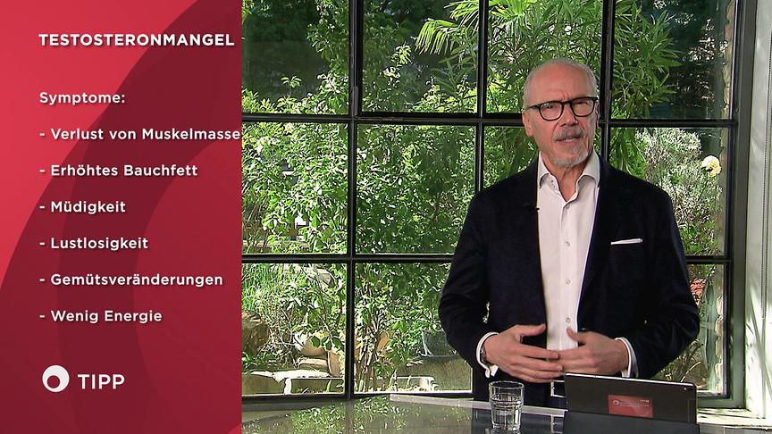 Prof. Dr. Siegfried Meryn steht an einem Tisch im Studio und erklärt die Symptome eines Testosteronmangels, die grafisch links eingeblendet sind.