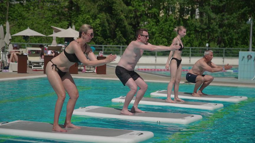 Mehrere Menschen in Badekleidung versuchen, auf schwimmenden Boards im Pool im Stehen eine Gleichgewichtsübung zu machen.