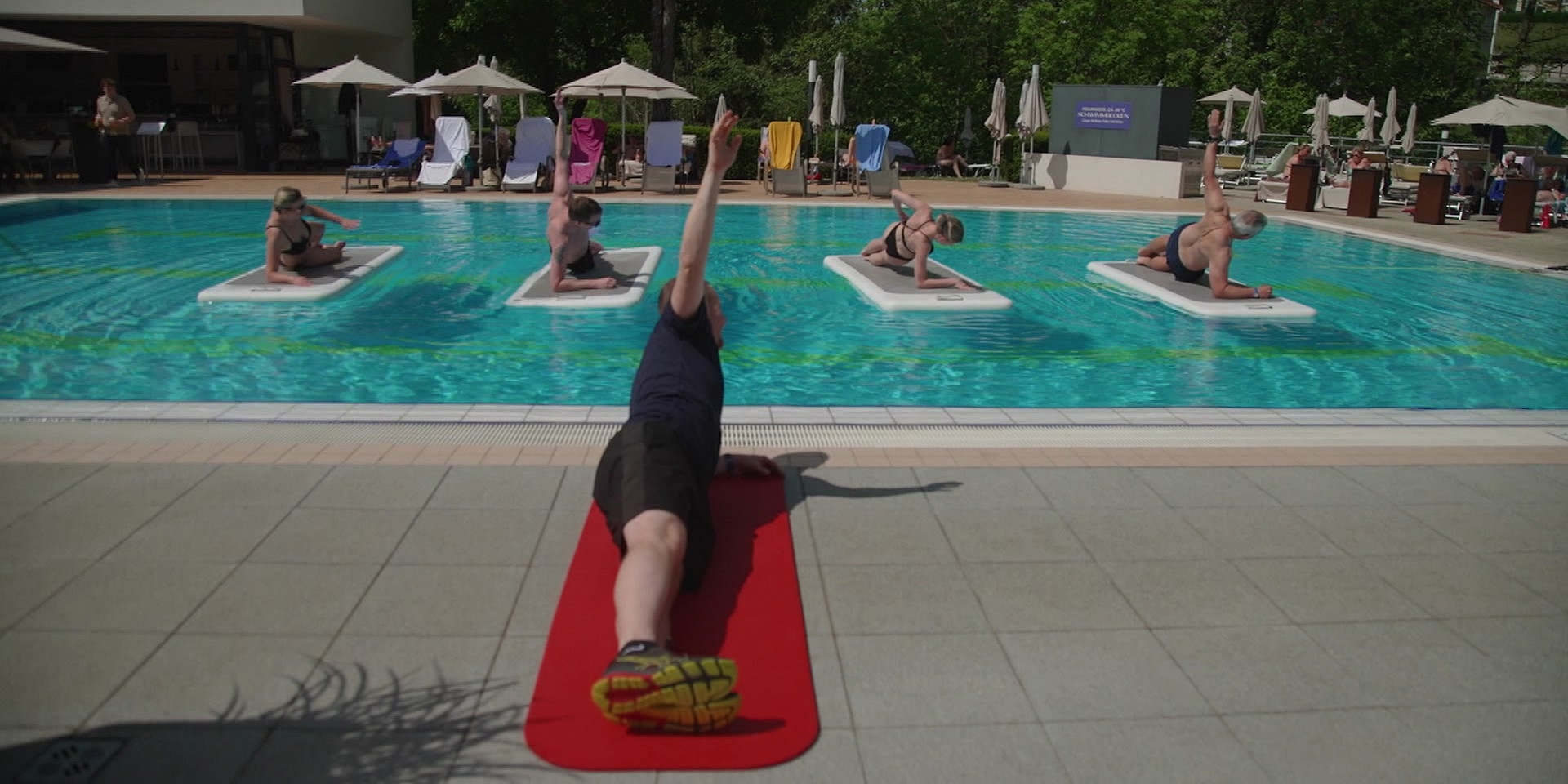 Ein Trainer führt am Beckenrand auf einer Matte eine Übung vor, während mehrere Menschen auf schwimmenden Wackelboard die Übung im Pool machen.