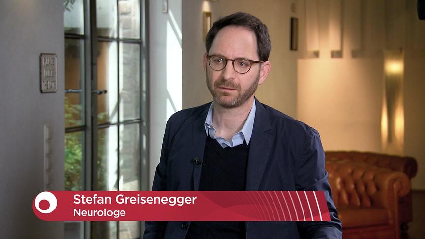 Stefan Greisenegger steht in einem Innenraum für ein TV‑Interview, eingeblendet ist eine Bauchbinde mit seinem Namen und der Berufsbezeichnung Neurologe.