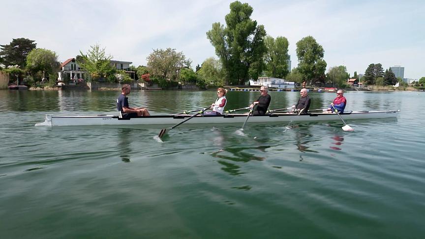 Ein Ruderboot mit mehreren Personen gleitet über einen ruhigen Fluss, im Hintergrund sind Bäume und Häuser am Ufer zu sehen.