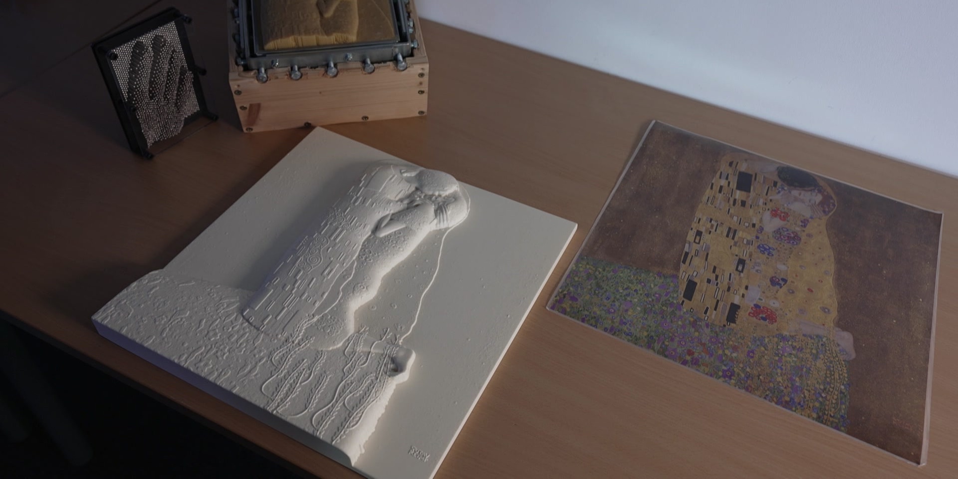 Auf einem Tisch liegen tastbare Reliefdarstellungen des Klimt-Gemäldes "Der Kuss" neben einer gedruckten Version, für sehbeeinträchtigtes Erleben.
