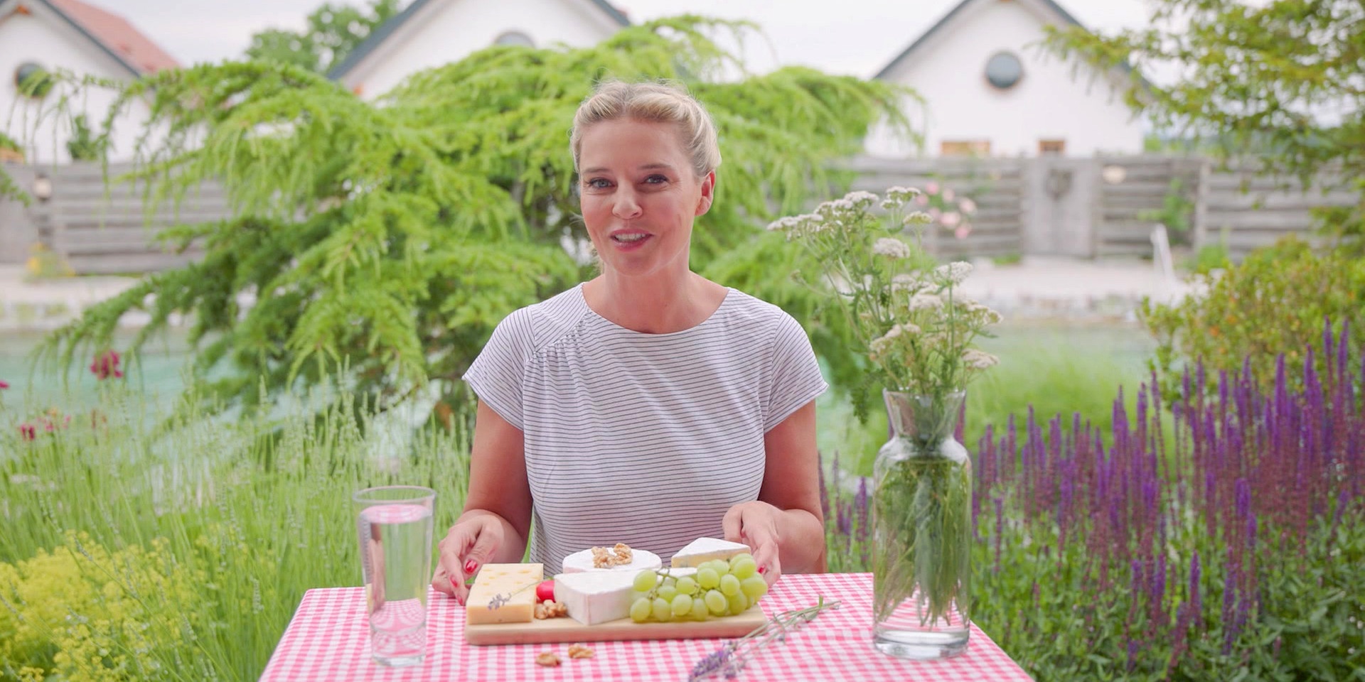 Christine Reiler sitzt an einem Gartentisch mit rot-weiß kariertem Tischtuch und einer Käseplatte mit Trauben, Nüssen, einem Glas Wasser und einer Vase mit Blumen vor einem grünen Gartenhintergrund.