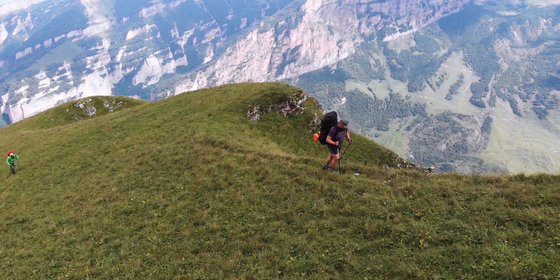 Zwei Wanderer steigen mit großen Rucksäcken über einen steilen, grasbewachsenen Bergrücken mit weitem Blick ins Tal.