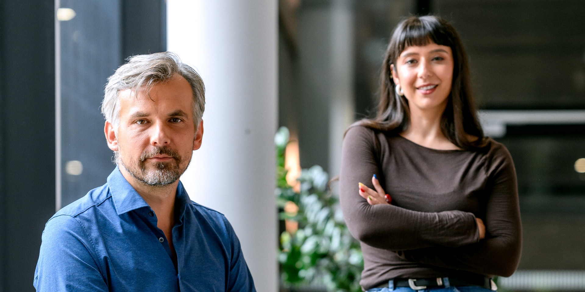 Moderator Tom Kamenar im blauen Hemd sitzt in einem modernen Innenraum, während Gestalterin Sandra Herbsthofer mit verschränkten Armen hinter ihm steht.