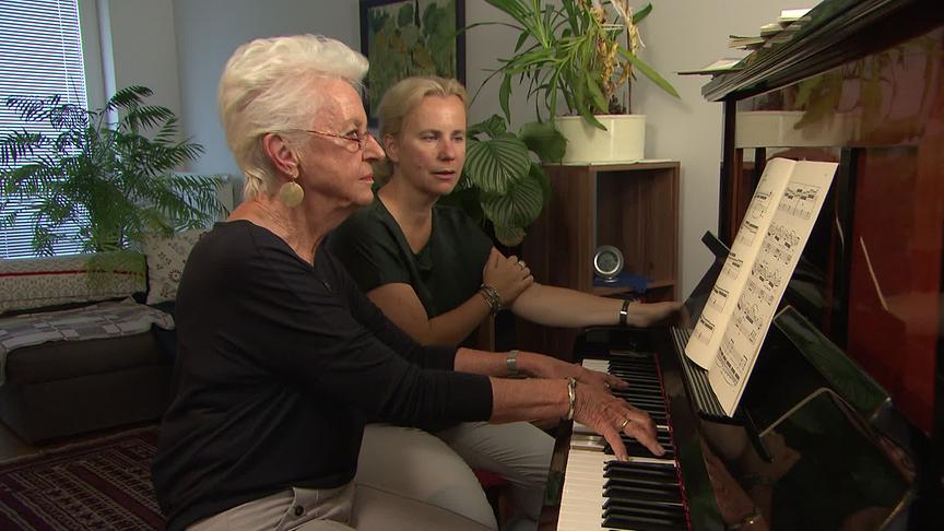 Musizieren im Alter: Eine ältere Frau spielt am Klavier in einem Wohnraum, neben ihr sitzt eine Frau, die ihr dabei zusieht.