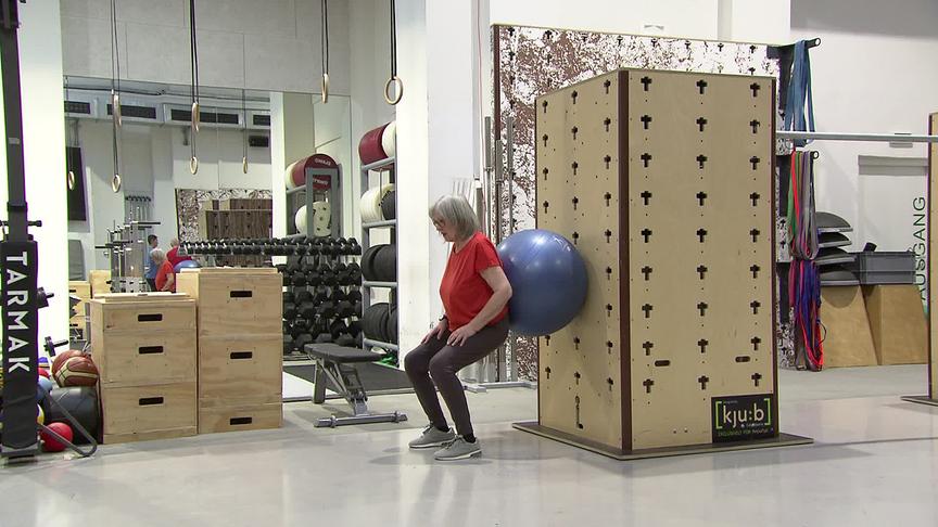 Eine ältere Frau trainiert im Fitnessstudio mit einem Gymnastikball Kniebeugen an der Wand.