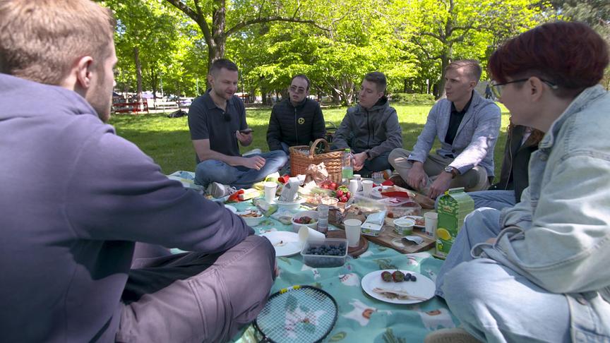Die Gruppe picknickt gemeinsam im Grünen, umgeben von Essen, Getränken und einem Korb.