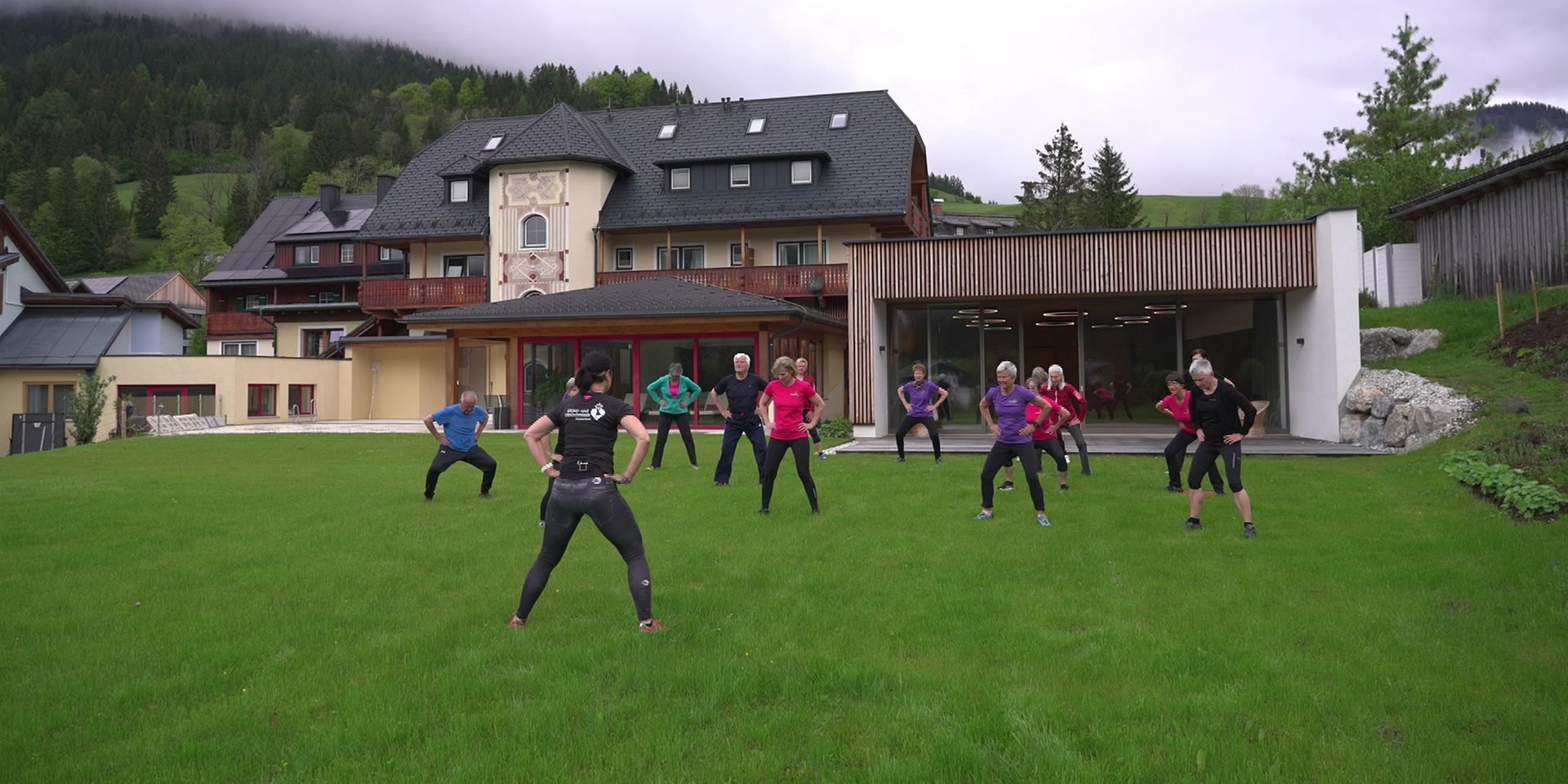 Eine Gruppe von Menschen macht gemeinsam Gymnastikübungen auf einer Wiese vor einem großen Haus in ländlicher Umgebung.