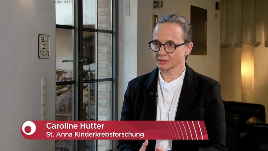 Eine Frau gibt ein Interview in einem modern eingerichteten Raum, eingeblendet ist ihr Name "Caroline Hutter, St. Anna Kinderkrebsforschung".