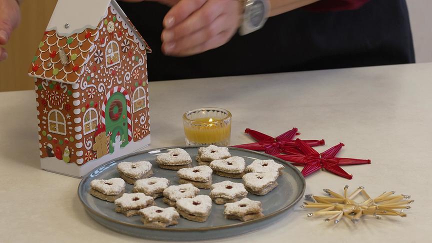Buchweizenkekse auf einem grauen Keramikteller nebeneinem Lebkuchenhaus, einer Kerze und roten Strohsternen.