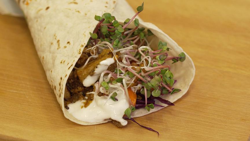 Ein gefüllter Wrap mit Wintergemüse, Sprossen und Sauce liegt auf einem Holzbrett.