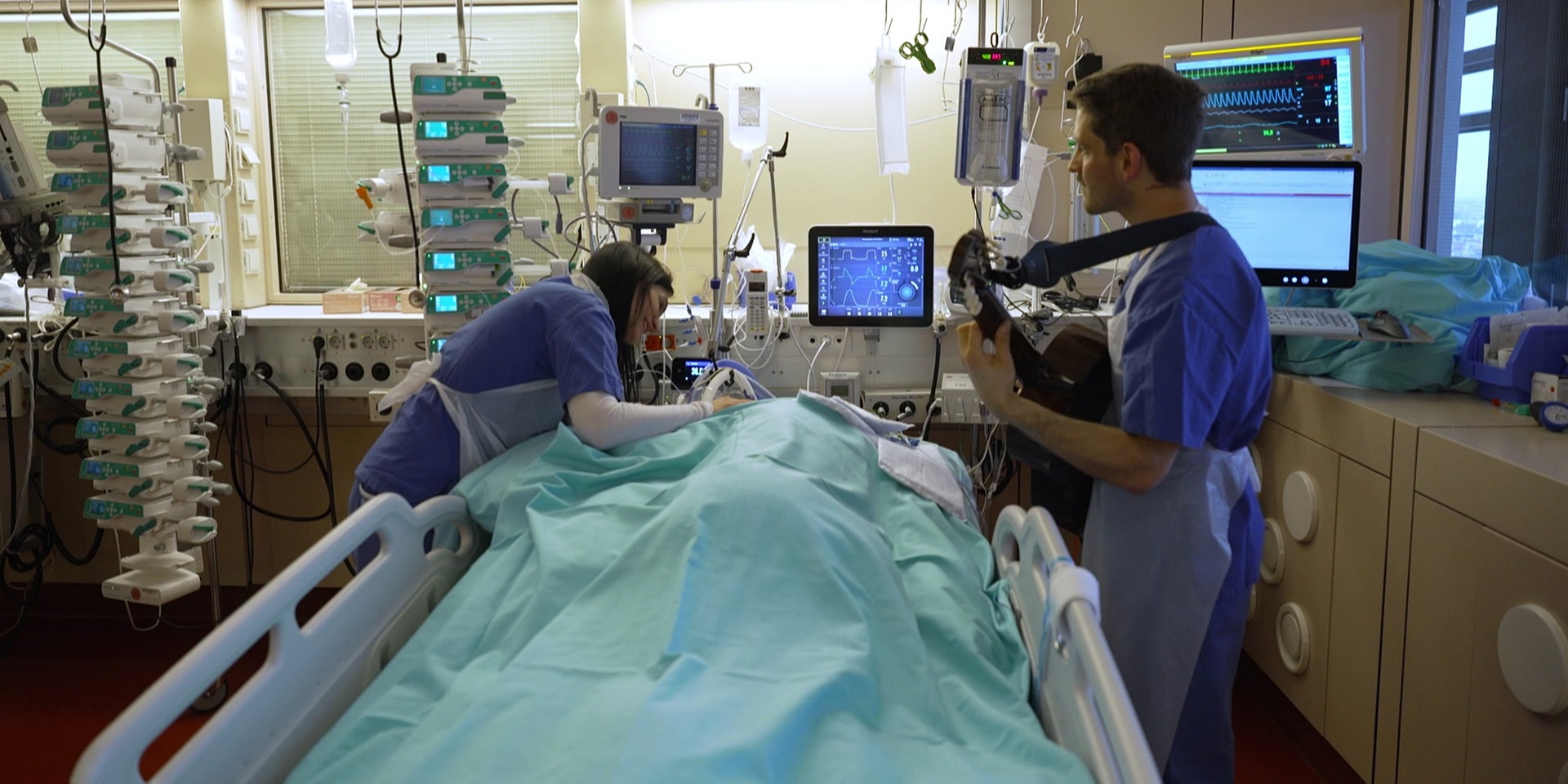 Zwei Pflegekräfte stehen bei einer intensivmedizinisch versorgten Person - eine davon spielt Gitarre.