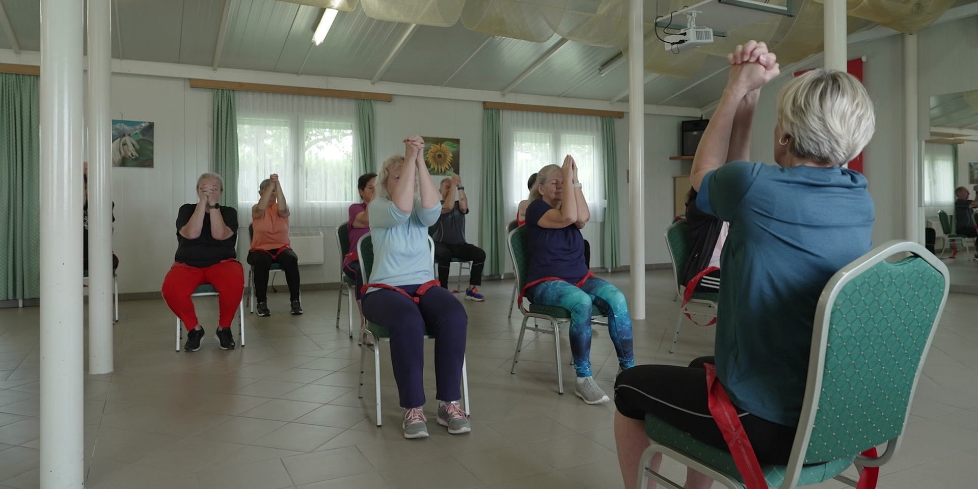 Eine Gruppe trainiert gemeinsam mit einer Übungsleiterin in einem Bewegungsraum auf Sesseln.