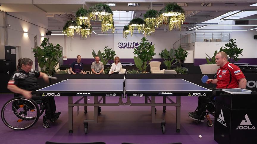 Henrik Andersson und eine Frau, beide im Rollstuhl. spielen Tischtennis in einer modern gestalteten Halle mit viel Begrünung.