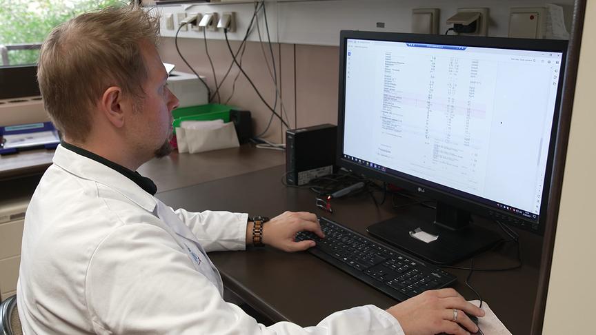 Ein Mann in einem weißen Laborkittel sitzt an einem Schreibtisch und arbeitet an einem Computer, auf dessen Bildschirm Daten von einem Bluttest angezeigt werden.