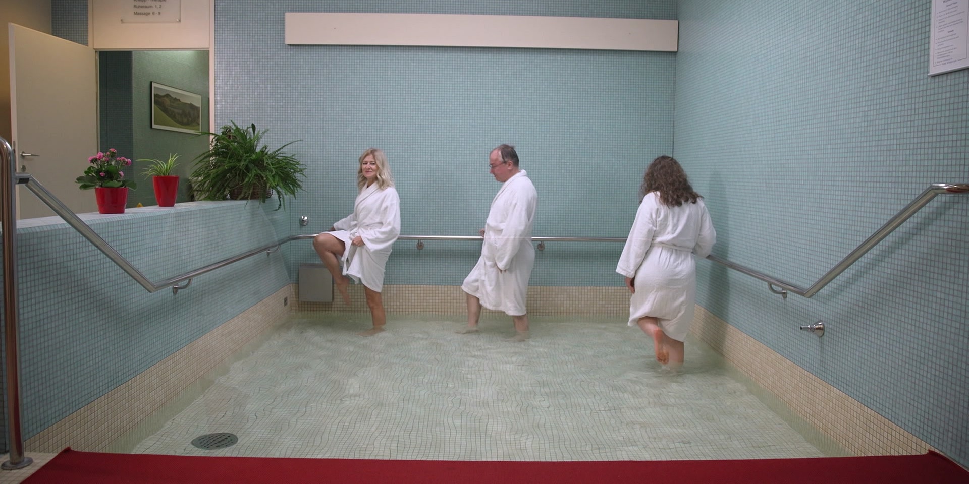 Zwei Frauen und ein Mann in Bademänteln führen eine Kneipp‑Wassertretübung in einem flachen Becken durch.