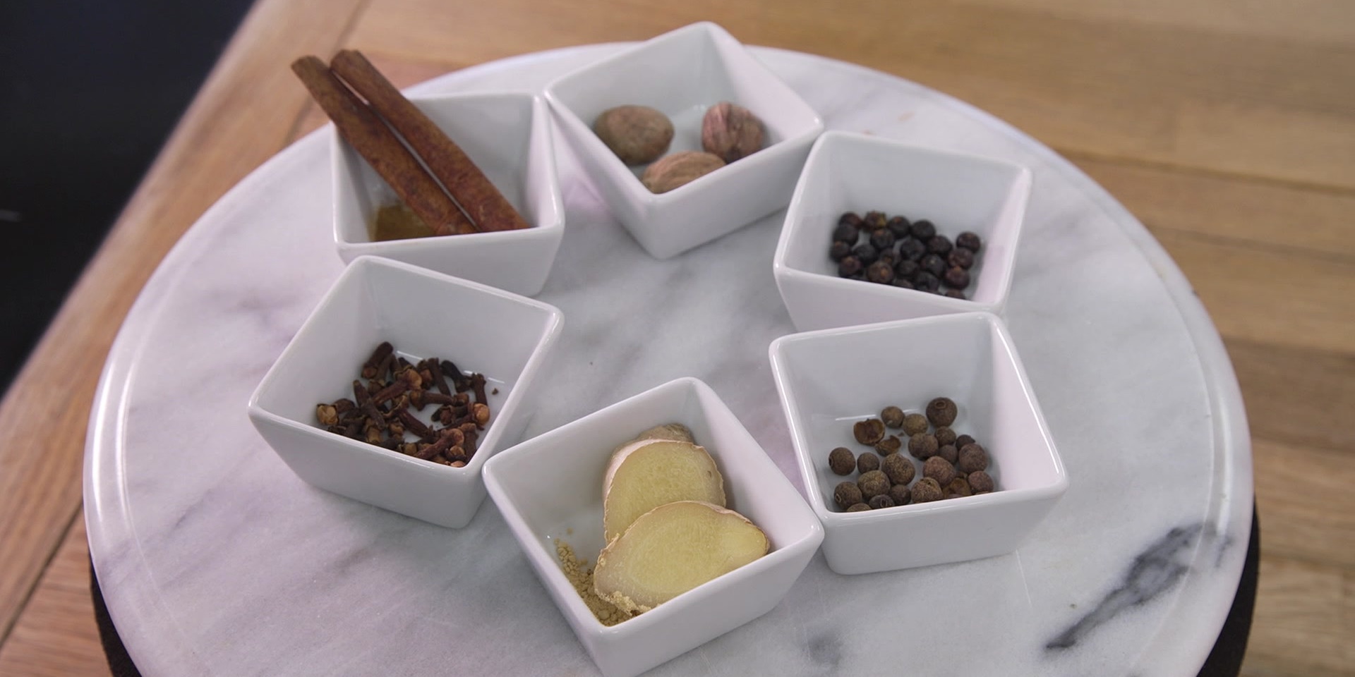 Verschiedene Gewürze wie Zimt, Muskat, Pfeffer und Ingwer liegen in kleinen weißen Schälchen auf einer runden Marmorplatte auf einem Holztisch.