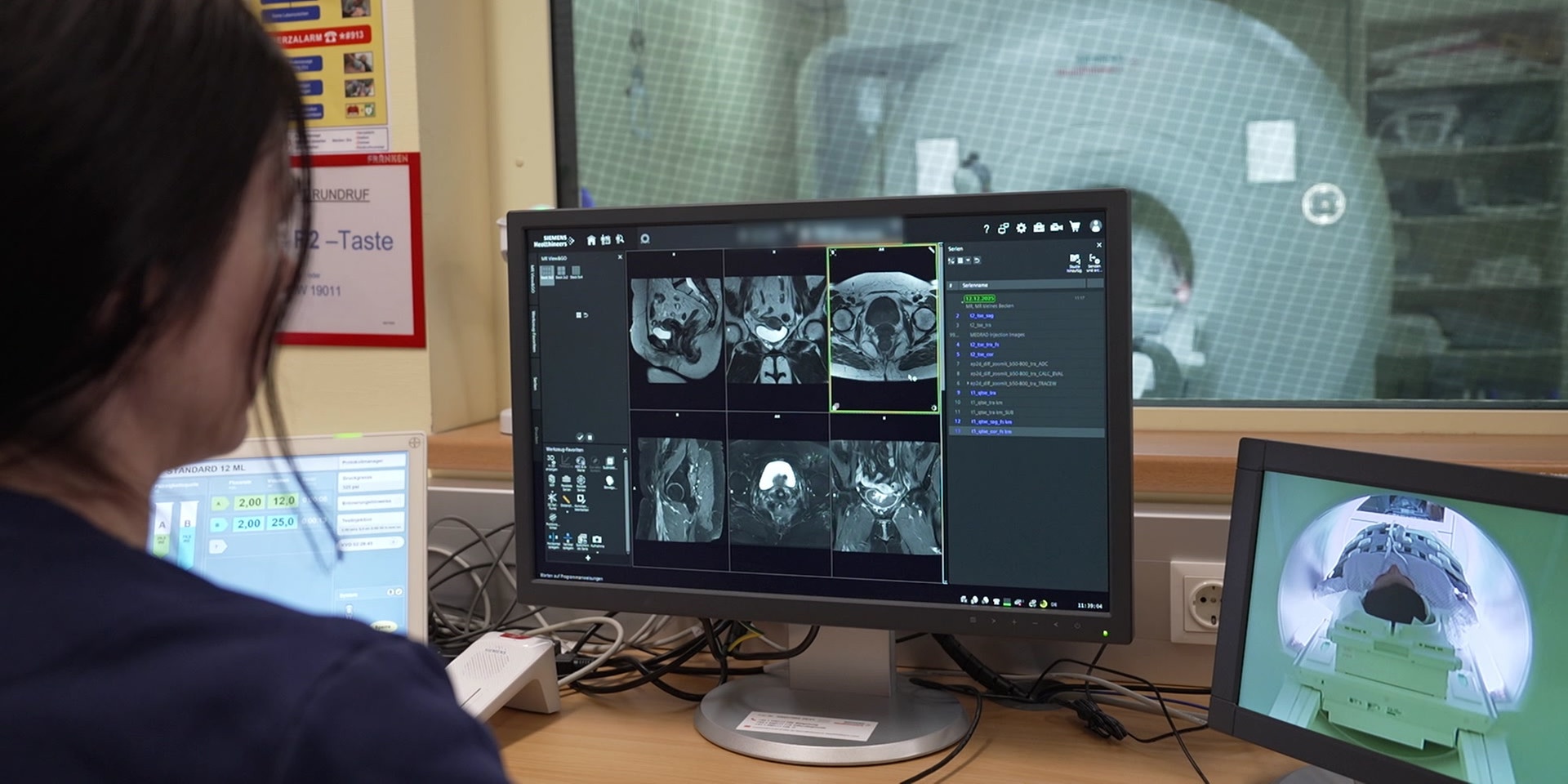 In einem medizinischen Kontrollraum betrachtet eine Frau mehrere Monitoren mit detaillierten MRT‑Aufnahmen, während hinter einer Glasscheibe das Magnetresonanztomografie‑Gerät mit einem liegenden Patient zu sehen ist.