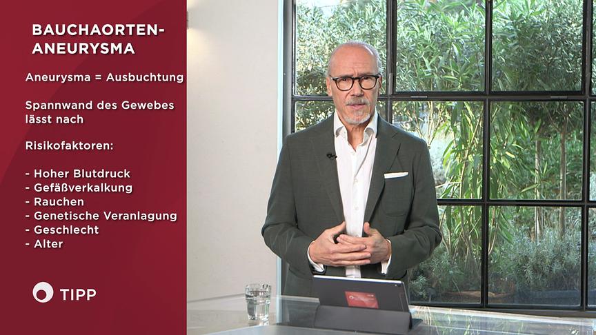 Prof. Dr. Siegfried Meryn  steht im Studio neben einer Infotafel zum Thema „Bauchaorten‑Aneurysma“ mit Definition und Risikofaktoren.