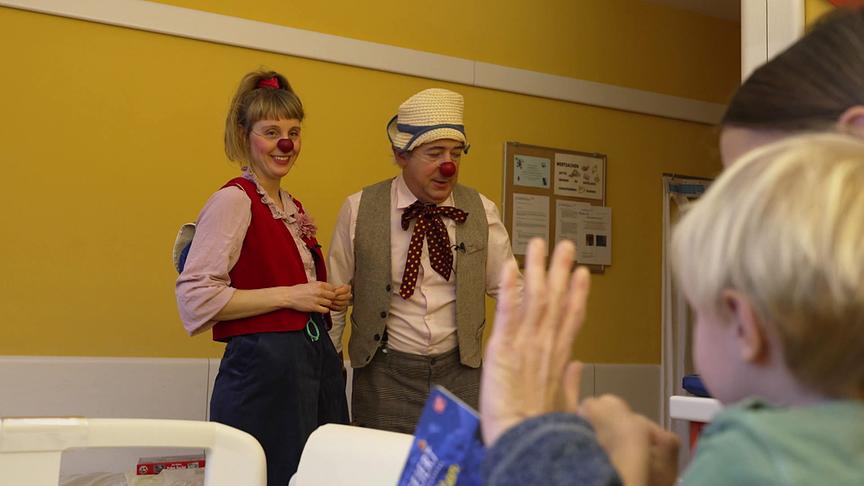 In einem Krankenzimmer stehen eine Frau und ein Mann als Clowns verkleidet mit Roten Nasen am Bett, während im Vordergrund ein Kind die Hand zum Winken hebt.