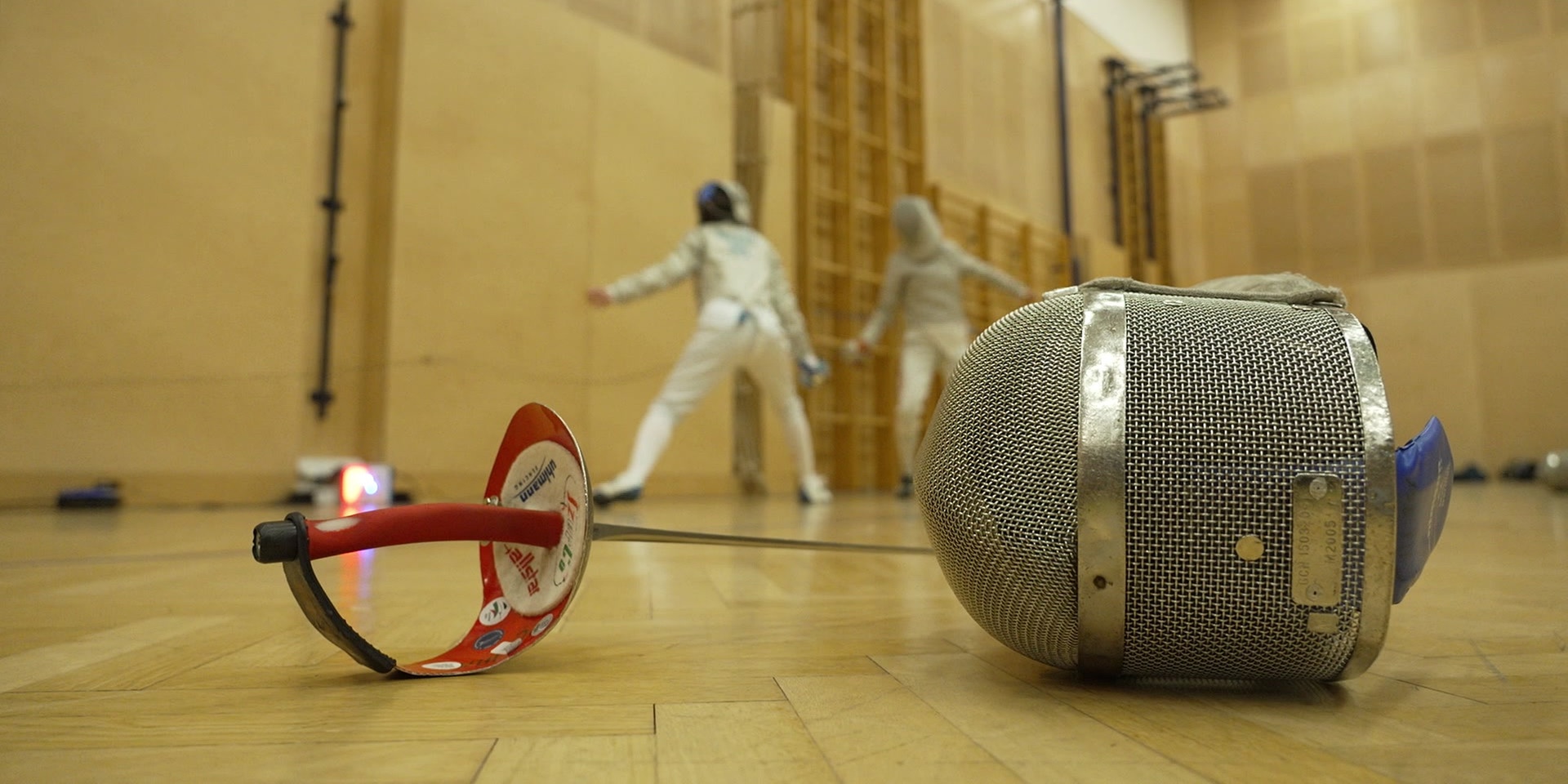Ein Degen und eine Fechtmaske liegen im Vordergrund auf dem Hallenboden, während im Hintergrund zwei Fechter in Aktion sind