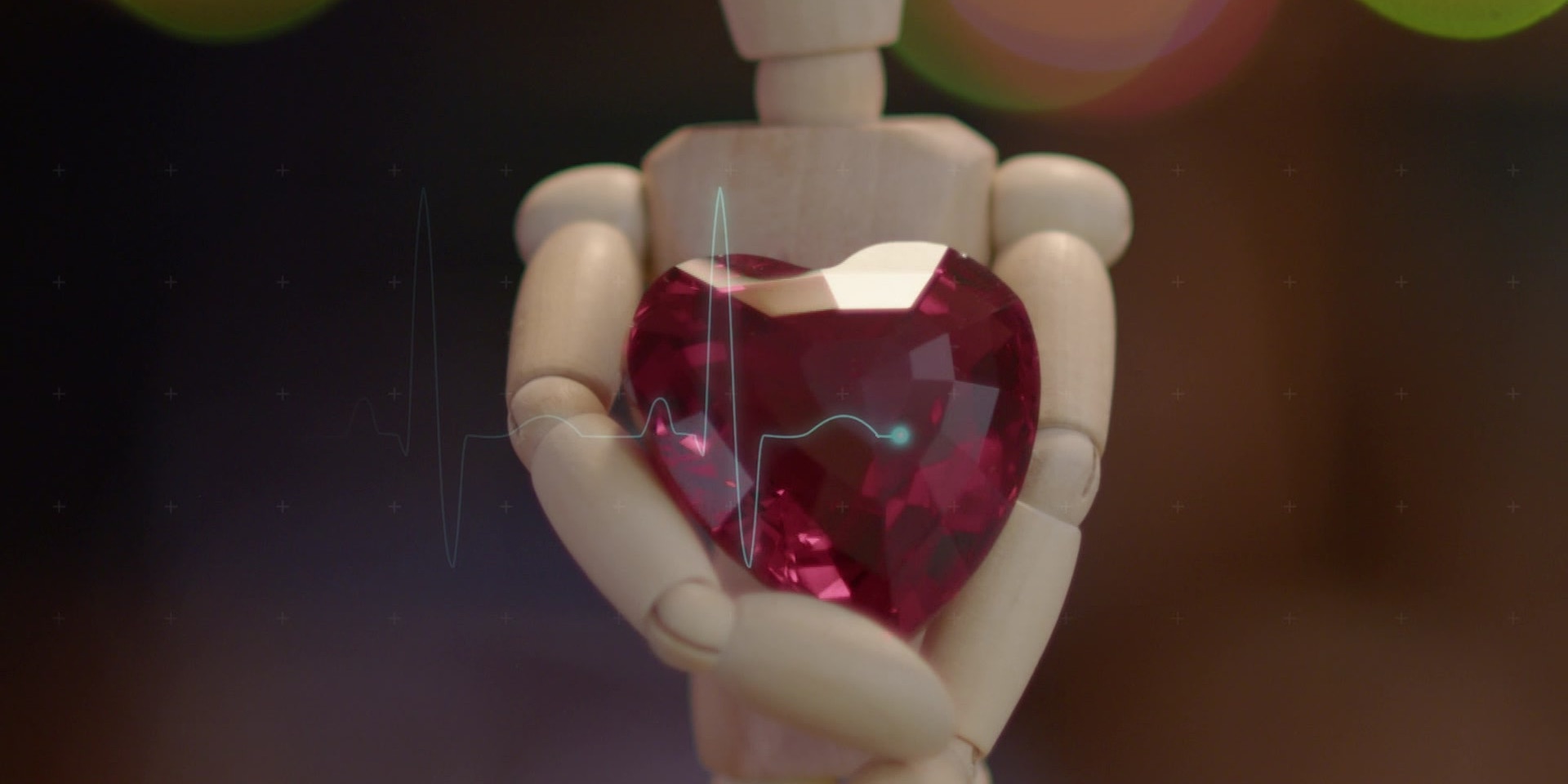 Eine Holzfigur hält ein facettiertes, rubinrotes Herz in den Händen, darüber liegt eine stilisierte Herzschlaglinie.