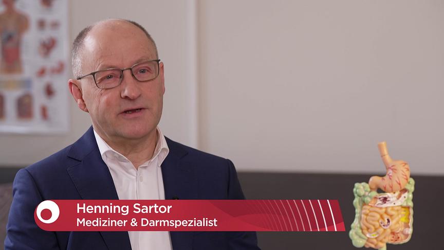Henning Sartor beim Interview in seiner Praxis. Eingeblendet ist eine Bauchbinde mit der Aufschrift „Henning Sartor – Mediziner & Darmspezialist“ neben einem Modell des Verdauungstrakts.