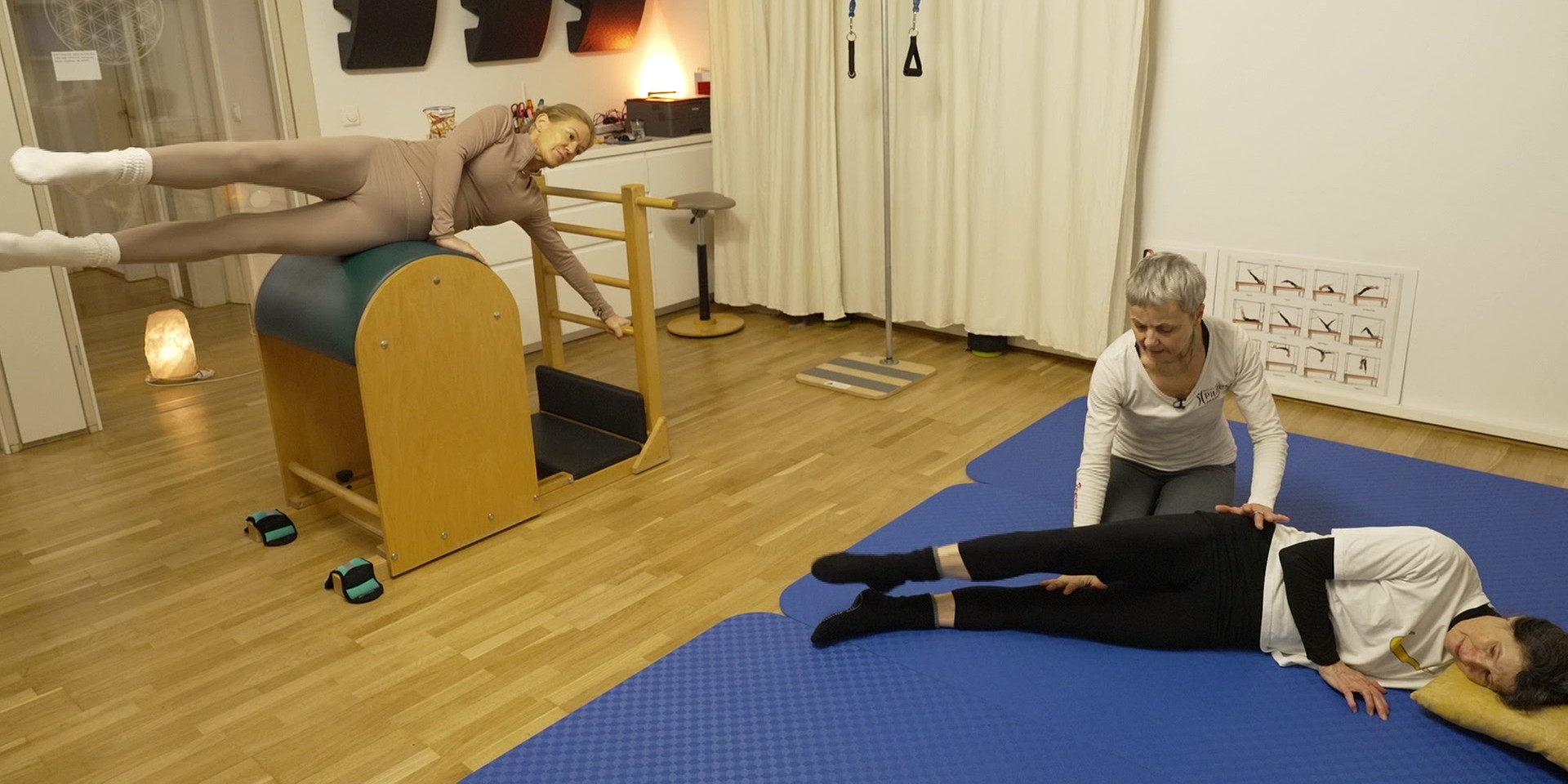 Eine Frau trainiert auf einem Pilates-Gerät, während eine andere auf einer Matte liegt und von einer Trainerin bei einer Übung unterstützt wird.