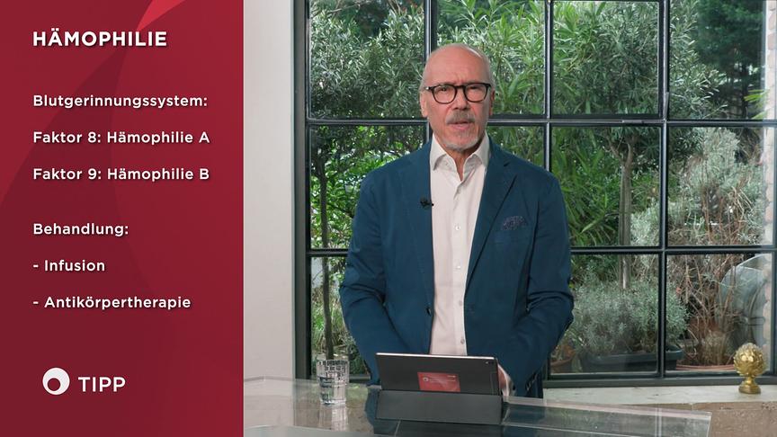 Prof. Dr. Siegfried Meryn steht an einem Tisch im Studio und erklärt Inhalte zu Hämophilie, die grafisch links eingeblendet sind.