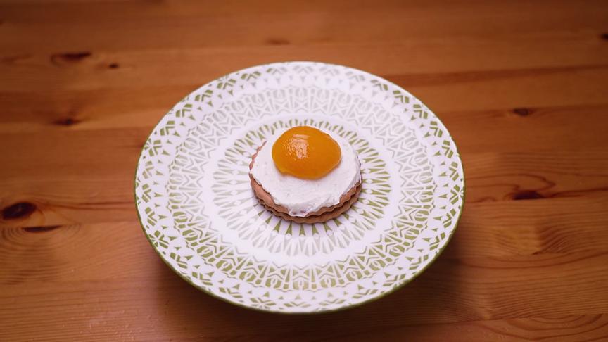 Ein rundes Keksgebäck ist mit einer weißen Topfencreme und einem Klecks Marillenmarmelade als süßes Spiegelei belegt.