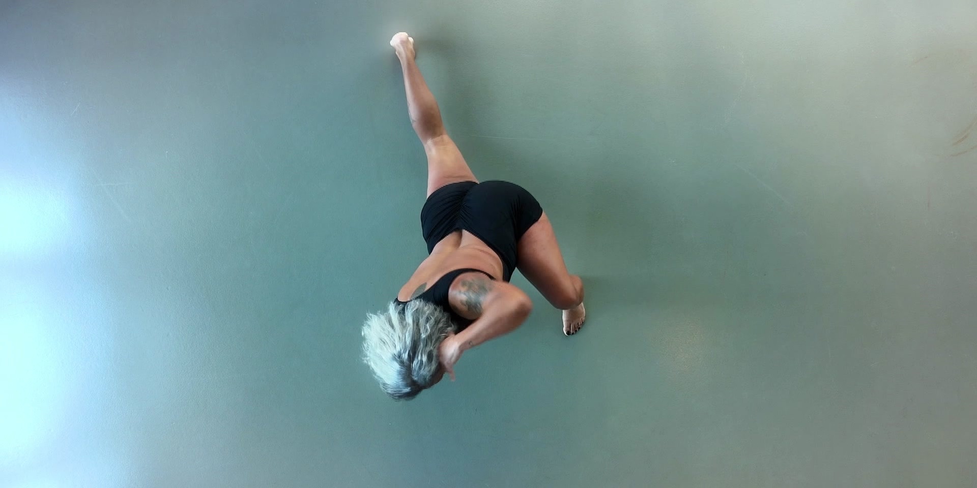 Eine Frau aus der Vogelperspektiv in schwarzer Sportkleidung, die aus der Vogelperspektive eine kraftvolle, tänzerische Bewegung auf einem glatten Boden ausführt.