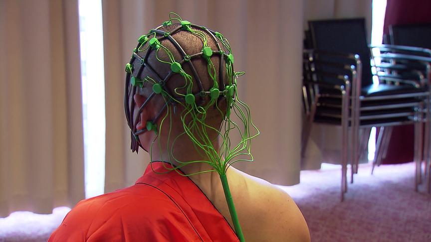 Ansicht des Hinterkopfs eines Shaolin-Mönchs in einer orangefarbenen Robe mit einer grünen EEG-Messhaube auf dem Kopf in einem Innenraum .