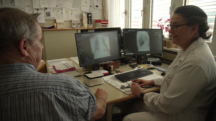 Eine Ärztin bespricht mit einem Patienten an einem Schreibtisch Röntgenbilder an zwei Monitoren.