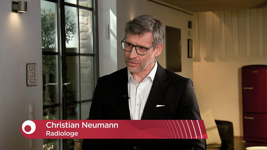 Radiologe Christian Neumann steht im Studio beim Interview, dessen Name und Berufszeichnung grafisch eingeblendet sind.