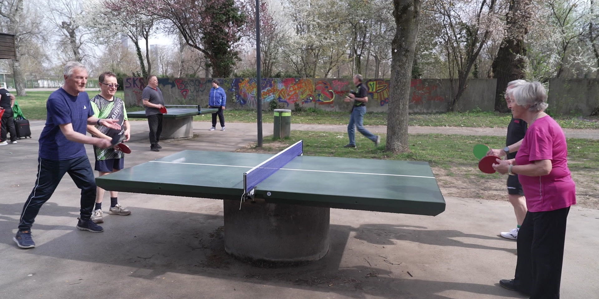 Mehrere ältere Menschen spielen Tischtennis an einer Outdoor-Platte in einem Park.