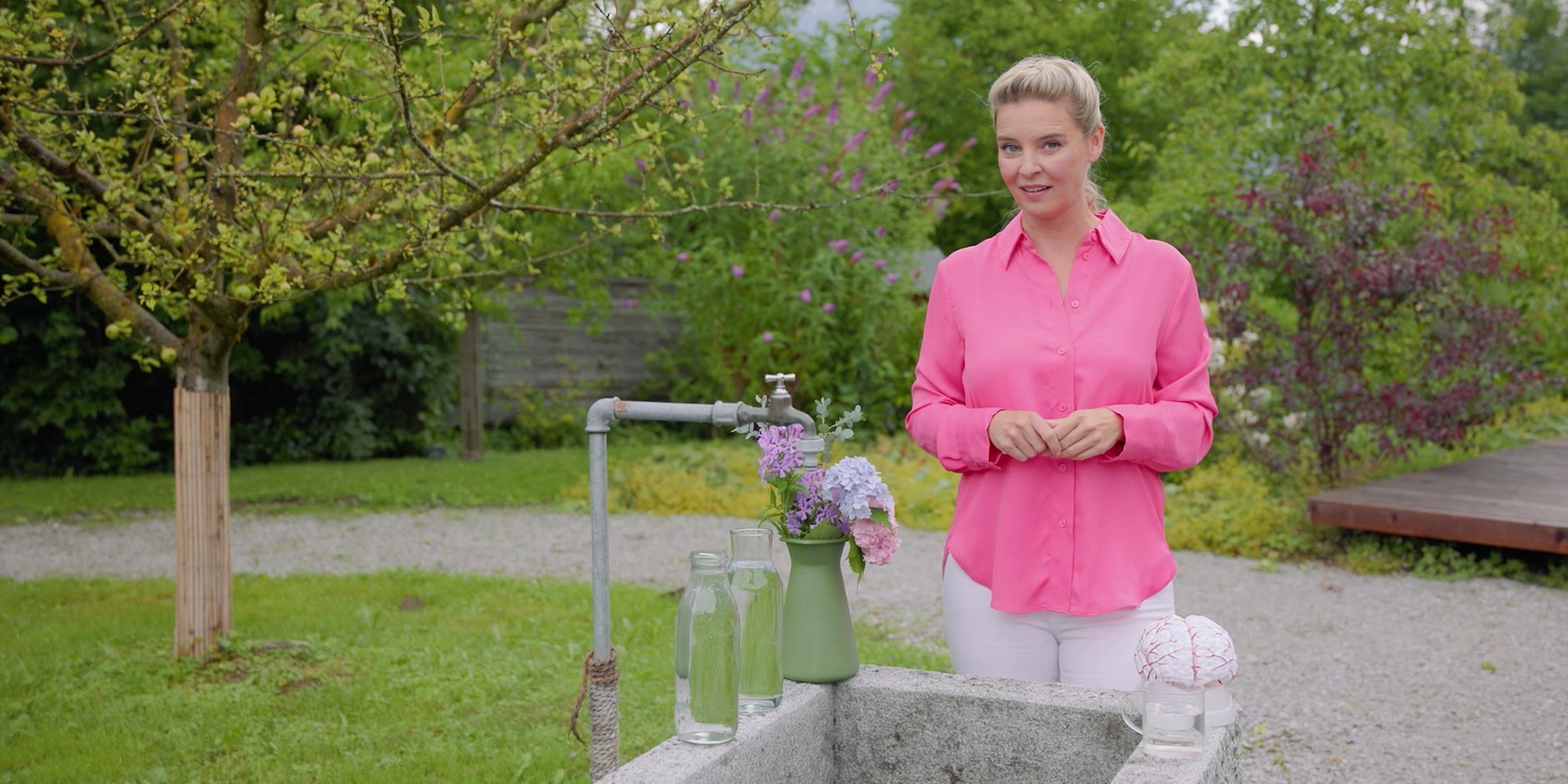 Dr. Christine Reiler in einer pinken Bluse steht in einem grünen Garten hinter einem steinernen Brunnen, auf dem Glasflaschen, Blumen und ein Modell eines Gehirns platziert sind.